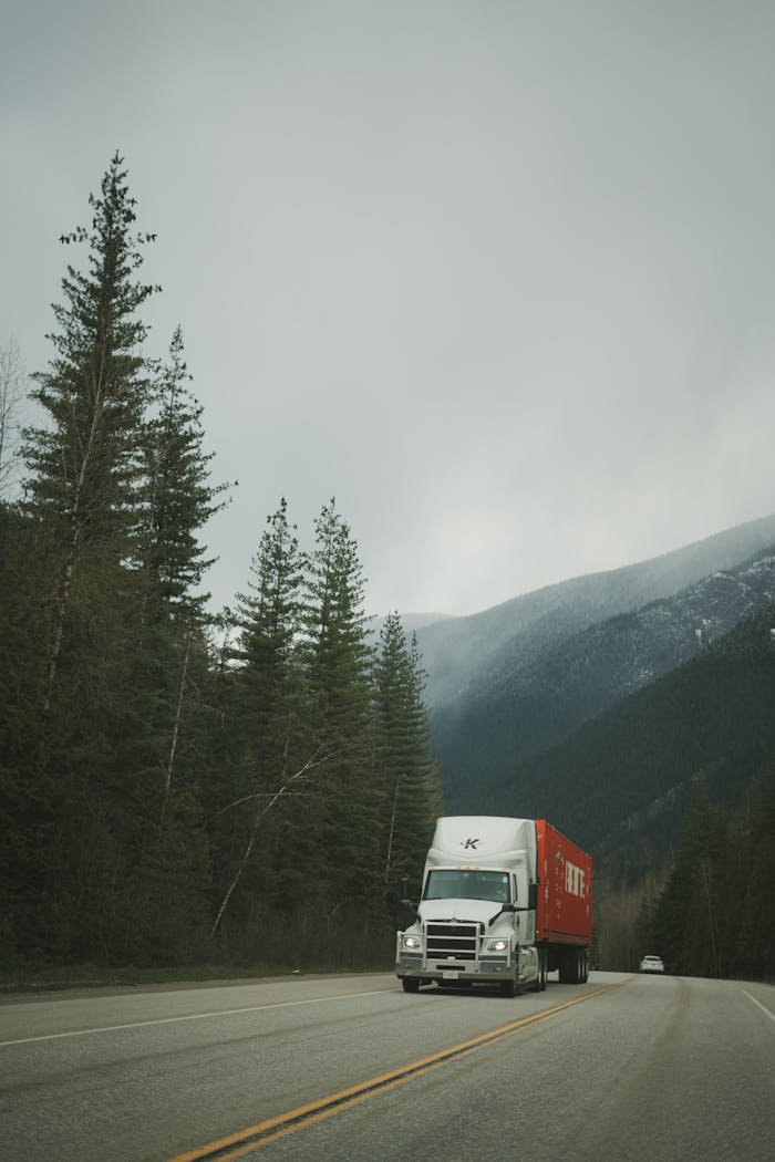A delivery truck drives along a scenic highway surrounded by mountains and evergreen trees.